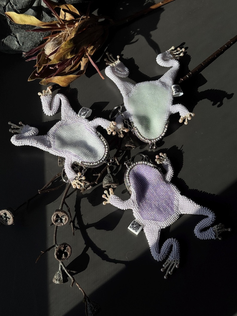 May include: Three beaded frog-shaped accessories with silver-tone accents. The frogs have white beaded bodies and light green or purple centers. Each frog has a small crown and tiny bells. The accessories are arranged on a dark surface.