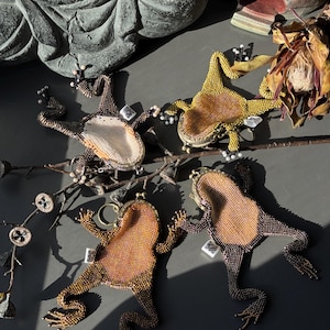 May include: Four beaded frog-shaped coin purses in shades of brown, gold, and green. Each frog has a unique color pattern and is embellished with small beads. The purses are arranged on a dark surface, with a branch and decorative elements.
