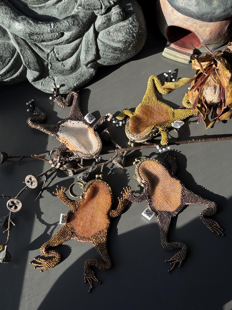 May include: Four beaded frog-shaped coin purses in shades of brown, gold, and green. Each frog has a unique color pattern and is embellished with small beads. The purses are arranged on a dark surface, with a branch and decorative elements.