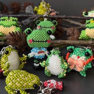 May include: A collection of beaded frog keychains in various colors, including green, white, and yellow. Some frogs have pink accents and bows. The keychains are attached to silver metal rings.