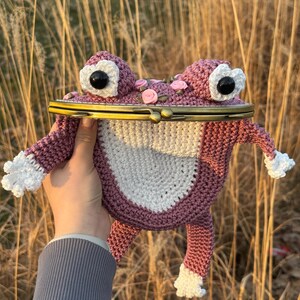 Könnte beinhalten: Handgefertigte, gehäkelte Frosch-Geldbörse in Mauve- und Weißtönen. Die Geldbörse hat große, weiß umrandete Augen mit schwarzen Zentren, kleine rosa Rosenakzente und einen Messingverschluss. Die Arme und Beine des Frosches sind ausgestreckt.