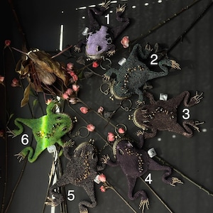 May include: A collection of beaded frog-shaped coin purses in various colors, including purple, green, and brown. Each purse features intricate beadwork, with gold-colored accents on the feet and a clasp closure. The purses are arranged on a dark surface with floral accents.