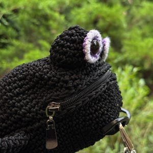 May include: Black crochet frog-shaped crossbody bag with a zipper closure and a detachable strap. The frog's eyes are detailed with white and purple accents. The bag is set against a blurred green background.