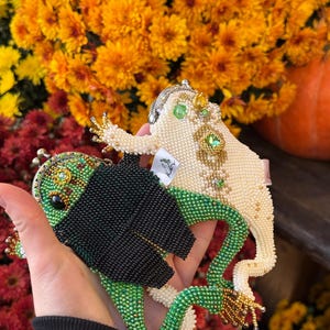 May include: Two beaded frog-shaped coin purses. One is green and black, the other is off-white with gold and green accents. The purses are held in front of a backdrop of orange and yellow flowers.