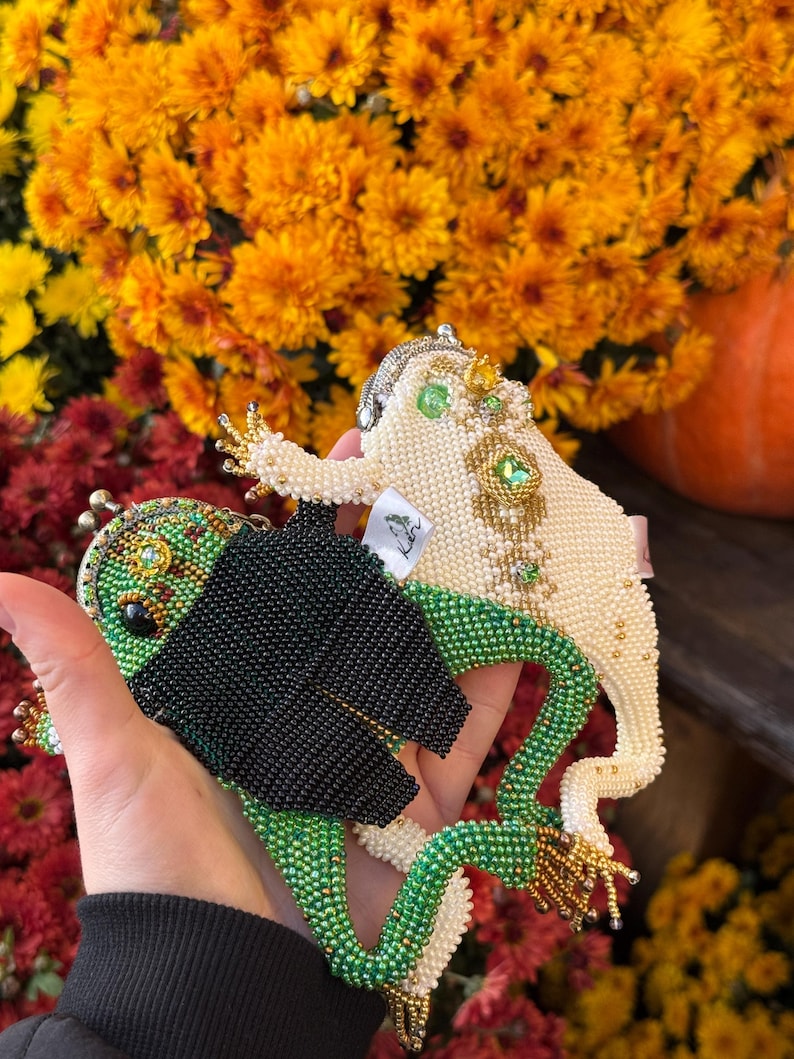 May include: Two beaded frog-shaped coin purses. One is green and black, the other is off-white with gold and green accents. The purses are held in front of a backdrop of orange and yellow flowers.