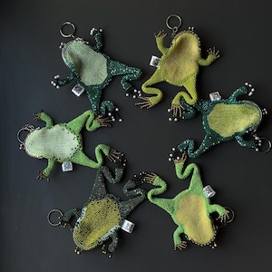 May include: Six beaded frog keychains in shades of green and yellow are arranged on a dark gray surface. Each frog has a metal clasp and a small tag. The frogs' bodies are covered in beads, with white bead accents.