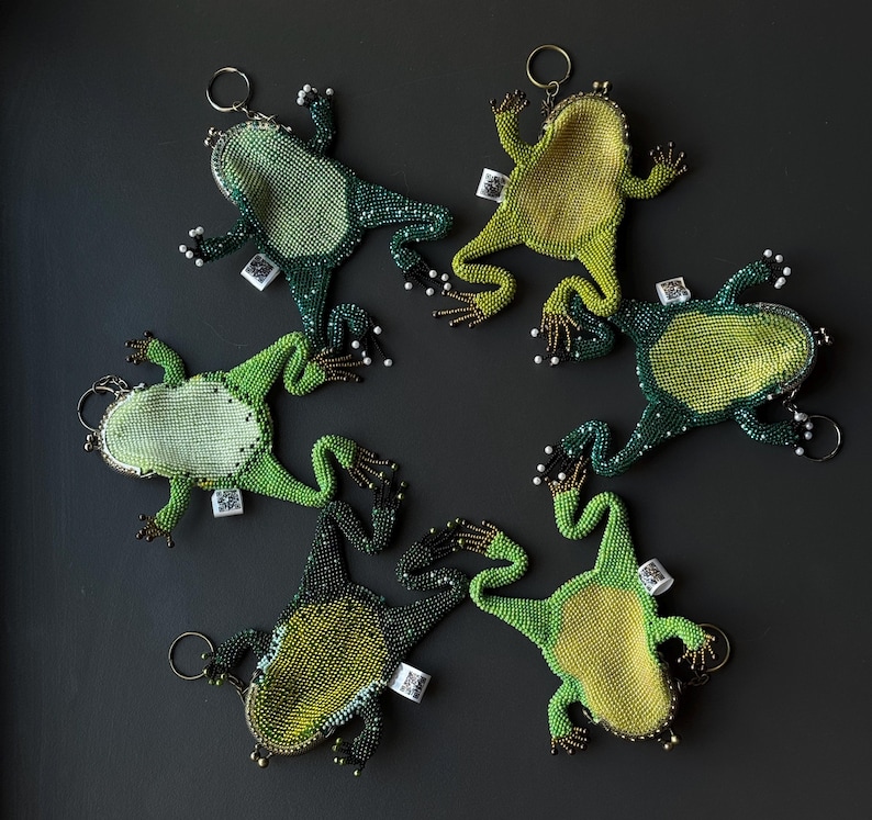 May include: Six beaded frog keychains in shades of green and yellow are arranged on a dark gray surface. Each frog has a metal clasp and a small tag. The frogs' bodies are covered in beads, with white bead accents.