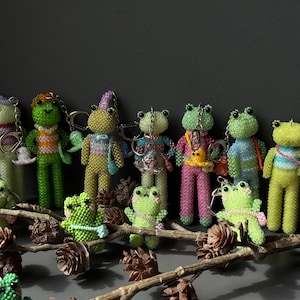 May include: A collection of beaded frog keychains in various colors, including green, yellow, and pink. The frog figures are adorned with accessories like hats and bags. They are arranged on a decorative branch with pinecones.