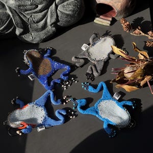 May include: Four beaded frog-shaped keychains in shades of blue, gray, and brown. The keychains have detailed beadwork, with some featuring white and red accents. The frogs are arranged on a dark surface, with a statue and decorative items in the background.