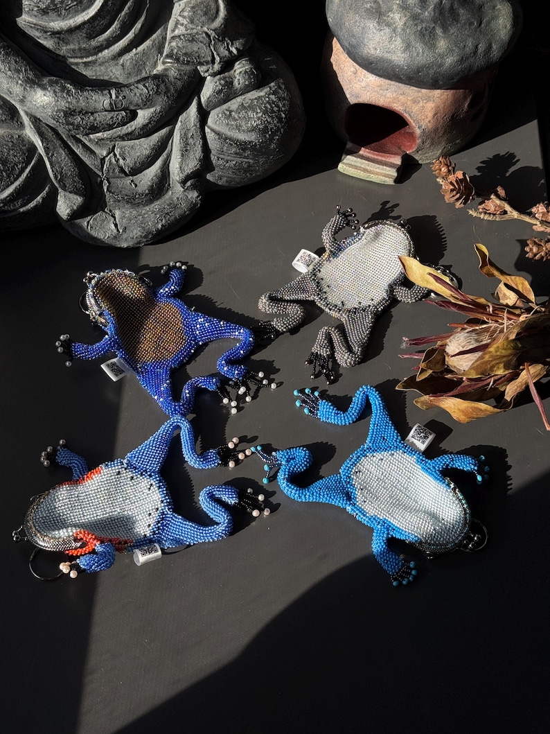 May include: Four beaded frog-shaped keychains in shades of blue, gray, and brown. The keychains have detailed beadwork, with some featuring white and red accents. The frogs are arranged on a dark surface, with a statue and decorative items in the background.