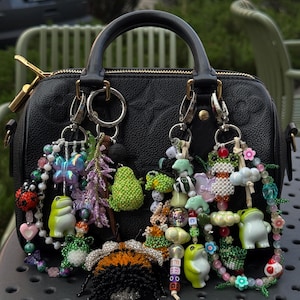 May include: A black leather handbag featuring a textured surface and two top handles, adorned with multiple colorful beaded keychains. The charms include various frog figures, a bumblebee, a butterfly, and floral beads attached to silver rings.