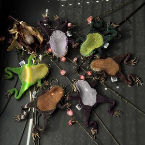 May include: A collection of beaded frog-shaped keychains in various colors, including green, brown, and purple. Each frog has a unique pattern and is adorned with gold-colored accents. The keychains are arranged on a dark surface with floral accents.
