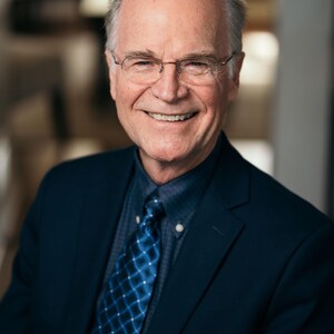 Puede incluir: Retrato de un hombre mayor sonriente con gafas, una chaqueta de traje azul marino, una corbata azul estampada y una camisa azul abotonada. El hombre tiene el pelo gris y una expresión cálida.
