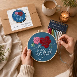 May include: An embroidery kit with a teal fabric hoop featuring embroidered pink and red roses and the words "Be Still" in white script. The kit includes a needle, thread, and a cardboard box with a matching design.