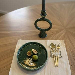 Könnte beinhalten: Tischarrangement mit einem grünen Keramikteller mit Feigen und Rosmarin, goldenem Besteck und einer grünen Kerze in einem dekorativen Halter. Ein beiger Leinen-Tischset und eine brennende Kerze vervollständigen das Arrangement.
