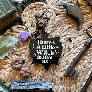 May include: A black keychain with a star and tassel, featuring the text "There's A Little Witch in all of us." The image includes two antique keys, small pumpkins, and a book with the word "Witch" on the spine.