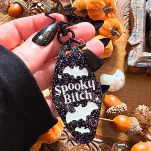 May include: A black and glittery keychain with the words "Spooky Bitch" and white bat silhouettes. The keychain has a black star-shaped top and a black tassel. The background includes miniature pumpkins and acorns.