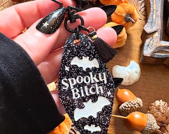 Puede incluir: Un llavero negro y brillante con las palabras "Spooky Bitch" y siluetas de murciélagos blancos. El llavero tiene una parte superior en forma de estrella negra y una borla negra. El fondo incluye mini calabazas y bellotas.
