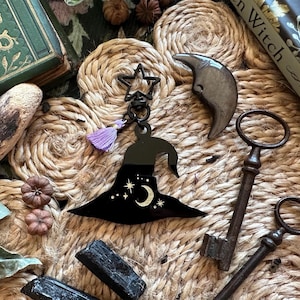May include: A black witch hat keychain with a star and moon design, a purple tassel, and a star-shaped clasp. The image also includes antique keys, a crescent moon, and small decorative pumpkins, all arranged on a woven surface.