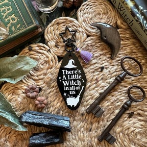 Pode incluir: Um porta-chaves preto com o texto "There's A Little Witch in all of us" e um gráfico de chapéu de bruxa. O porta-chaves tem um pendão roxo e um fecho em forma de estrela. Também são mostradas duas chaves antigas, cristais e objetos decorativos.