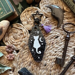 May include: A black and white cat-shaped keyring with a star-shaped clasp and a purple tassel. The keyring features a white cat and crescent moon design against a starry background. Other items include keys, a claw, and a book.