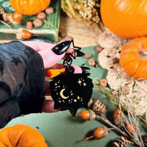May include: Black cauldron-shaped keychain with a crescent moon and star design. The keychain has a small orange tassel and a star-shaped clasp. The background includes pumpkins, acorns, and other fall-themed decorations.