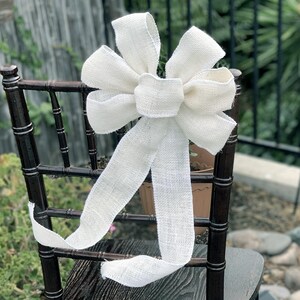 May include: A large, cream-colored burlap bow is tied to the back of a dark brown wooden chair. The bow has multiple loops and long, flowing tails. The chair is outdoors, with greenery visible in the background.