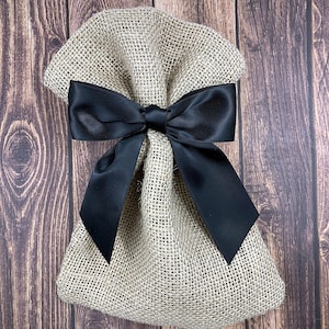 May include: A burlap gift bag tied with a large black satin bow. The bag is made of a coarse, woven fabric, and the bow is centered on the front. The background is a dark wood grain.