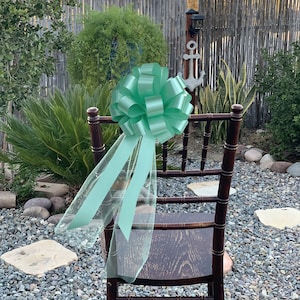 May include: A mint green satin bow with a sheer tulle tail attached to a brown wooden chair. The chair is in an outdoor setting with rocks and greenery.