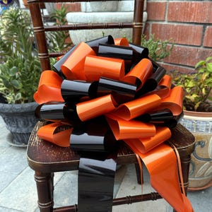 May include: A large decorative bow made of glossy black and orange ribbon. The bow is placed on a vintage wooden chair. The bow's vibrant colors and design make it suitable for festive occasions.