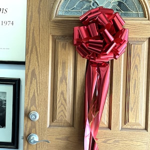 May include: A large, red, shiny bow is tied to a wooden door. The bow is made of multiple loops of ribbon and is tied in a knot at the top. The bow is hanging from the door handle.