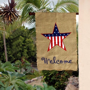 May include: A burlap welcome flag with a red, white, and blue star in the center. The star is in the style of the American flag. The text "Welcome" is embroidered in blue below the star.