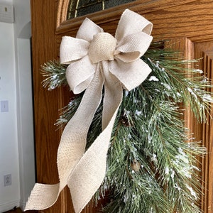 May include: A beige burlap bow with long tails is tied to a green pine garland with white faux snow. The garland is hanging on a wooden door.