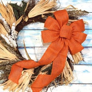 May include: A rustic fall wreath with a large orange burlap bow. The wreath is made of dried grasses and other natural materials.