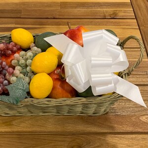 May include: A wicker basket filled with fruit, including grapes, lemons, apples, and avocados. A large white satin bow is tied around the top of the basket.
