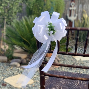 May include: White satin chair sash with a large bow and a small cluster of white flowers and green leaves.