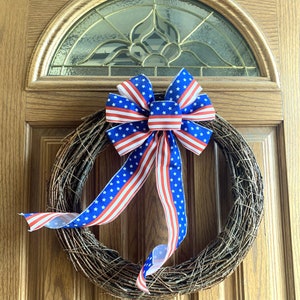 May include: A grapevine wreath adorned with a patriotic bow. The bow features a blue background with white stars and red and white stripes. The wreath is hanging on a wooden door with a decorative glass panel.