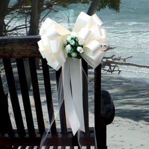 May include: A white satin bow with a tulle tail and a cluster of white flowers, tied to a wooden chair.