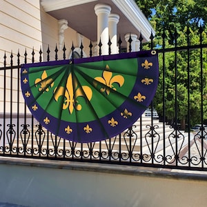 May include: A purple and green Mardi Gras fan with gold fleur-de-lis designs. The fan is hanging on a black metal fence.