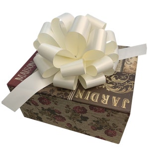 May include: A white satin bow tied on top of a gift box with a brown and gold floral design. The box has the words "JARDIN" and "MAISON" printed on it.