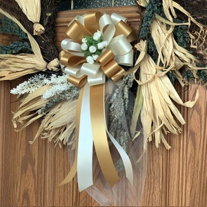 May include: A gold and white bow with a white tulle overlay and white flowers. The bow is attached to a wreath made of dried corn husks.