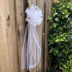 May include: A white tulle bow with a white satin ribbon tied around a wooden fence post.