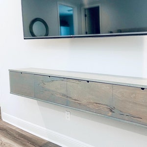 May include: A long, light gray wooden media console with four drawers. The console is mounted to the wall and has a modern, minimalist design.