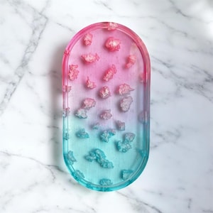 May include: An oval-shaped tray featuring a smooth gradient transition from pink at the top to blue at the bottom. The surface is embedded with numerous small, irregular, translucent crystalline fragments that are scattered throughout the tray.