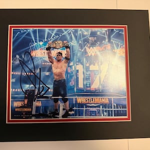 Peut inclure: Photographie encadrée d'un lutteur tenant une ceinture de championnat au-dessus de sa tête dans un ring. L'image est signée au marqueur noir. L'arrière-plan présente les mots "Wrestlemania" et le logo WWE.