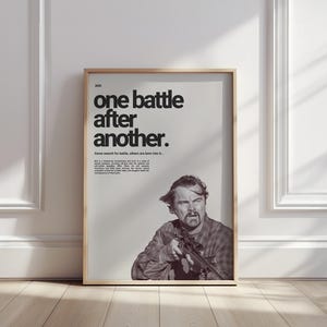 May include: Framed poster with the text "one battle after another." The poster features a black and white image of a man holding a rifle. The poster is in a wooden frame and leans against a white wall.