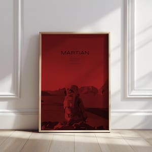 May include: A red movie poster with a white frame. The poster features the title "The Martian" in black text and a silhouette of an astronaut in a spacesuit standing on a red, rocky surface.