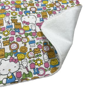 Puede incluir: Una toalla de cocina de tela blanca con un estampado colorido de caras sonrientes, flores y un personaje de Hello Kitty. La toalla tiene un respaldo de felpa blanca.