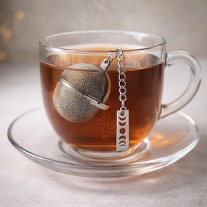 May include: A clear glass teacup filled with tea, with a stainless steel tea infuser. The infuser is a mesh ball with a chain and a charm featuring moon phases. The cup sits on a clear saucer.