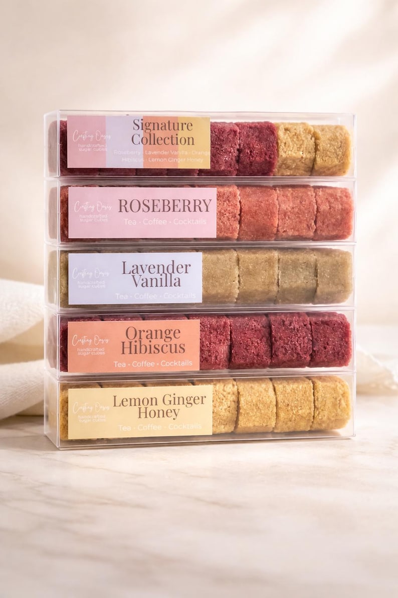 Op de afbeelding: Een stapel van vijf transparante dozen, elk met handgemaakte suikerklontjes. Smaakvariaties zijn onder meer Roseberry, Lavender Vanilla en Orange Hibiscus. De dozen zijn gelabeld met het "Crafting Drive" logo en de smaaknamen.